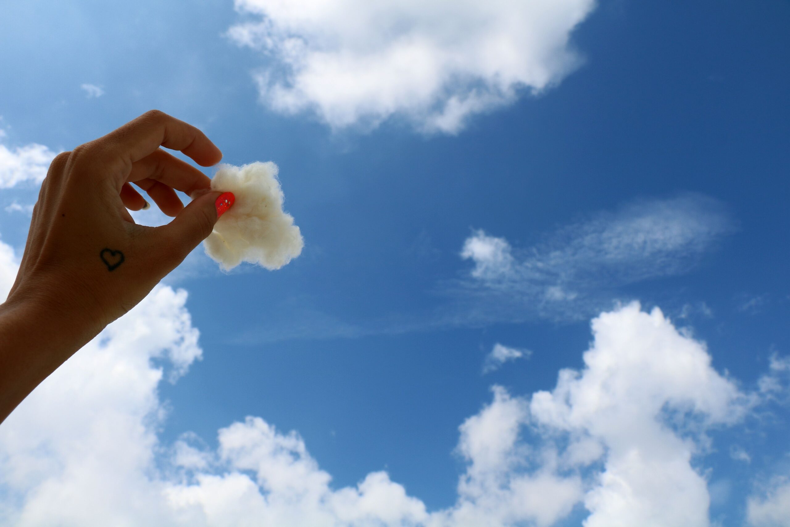 A hand with a heart tattoo holds cotton resembling a cloud in a clear blue sky.